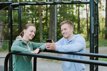 Obraz premium Caucasian young woman and Caucasian young man standing outdoors near metal bars, smiling and looking at smartphone together, engaging in conversation in park setting