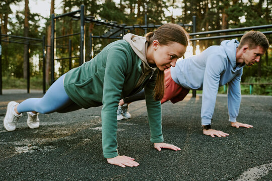 Caucasian young woman and Caucasian young man exercising outdoors performing push ups on asphalt surface in park setting, both focused on workout, athletic activity in progress