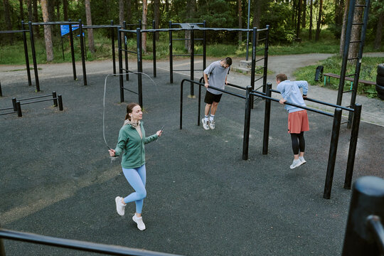 Caucasian young woman skipping rope while Caucasian teenage boy and Caucasian teenage girl exercising on outdoor fitness bars in park setting, all focused on physical activity