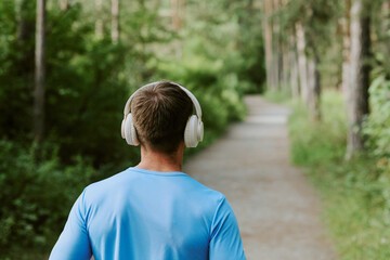 Caucasian young adult man walking on forest path wearing headphones, listening to music or podcast, enjoying outdoor activity, back view with green trees and nature surrounding