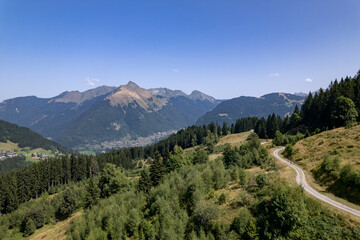 Aerial mountainous landscape with meandering path through lush green French Alps valley tourist destination winter sports near Les Gets village with rocky mountain top against a clear blue sky. 