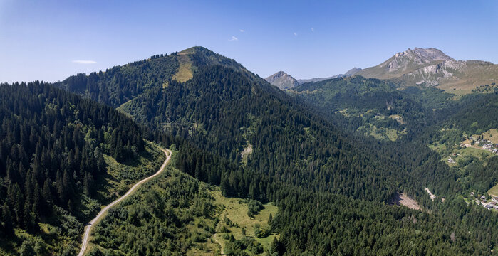 Aerial mountainous landscape with meandering path through lush green French Alps valley tourist destination winter sports near Les Gets village with rocky mountain top against a clear blue sky. 