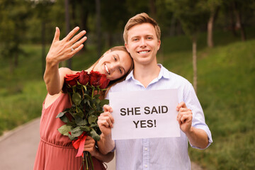 Smiling man holding sheet of paper with text She Said Yes and his happy beloved with bouquet of...