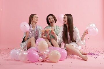 Pajama party. Happy friends with tinsel and balloons clinking glasses on pink background