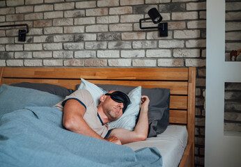middle-aged bald man Portrait with short beard wearing black sleeping mask lying comfortably in bed...