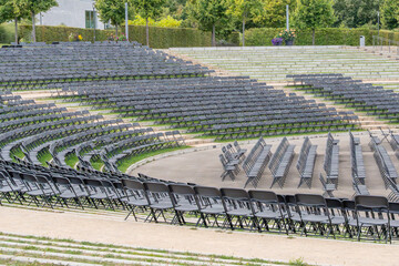 An empty outdoor amphitheater with rows of black folding chairs arranged in arcs. The seats await...