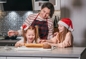 Cute little girls in red Santa hats with mother making homemade dough Christmas gingerbread cookies...