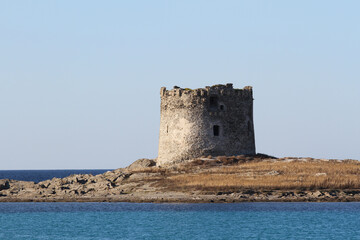 Close-up view of an ancient coastal tower on a rocky islet in Sardinia under a clear blue sky. The historic structure stands as a sentinel over the Mediterranean Sea.