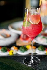 Red cocktail with mint and lime garnish in a wine glass, served at a fine dining table with assorted appetizers in the background. Elegant, festive, and refreshing atmosphere