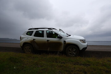 White SUV parked by a gravel coastal road in Iceland, with mud stains on the side and dramatic cloudy sky above the sea in the background