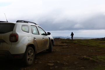 sUV parked on a rugged coastal road in Iceland, with a traveler holding a camera and dramatic...