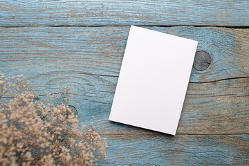 Blank white card placed on rustic wooden surface with blue paint, surrounded by delicate dried...