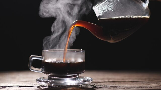 Hot coffee being poured into a clear glass cup with steam rising against a dark background, symbolizing warmth, freshness, and energy. Concept of hot coffee drink.