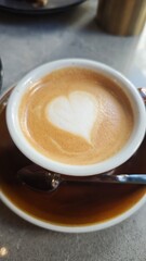 Cup of cappuccino with heart-shaped latte art on a saucer and spoon on a cafe table. Concept of morning ritual, warmth and cozy coffee break moment