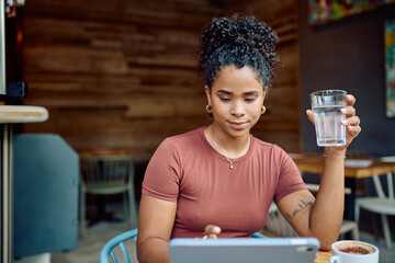 Young woman sitting in a cafe, enjoying a glass of water while using a digital tablet for browsing...