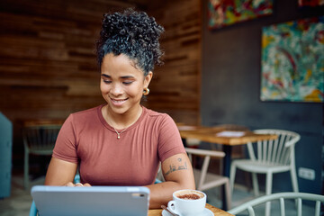 Smiling young woman enjoying a cappuccino while browsing on a digital tablet at a cozy cafe table, embracing a modern lifestyle