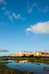 Old town of Essaouira, Morocco, seen at low tide in the evening light. Reflections of white buildings and city walls in tidal pools with seaweed and dramatic sky