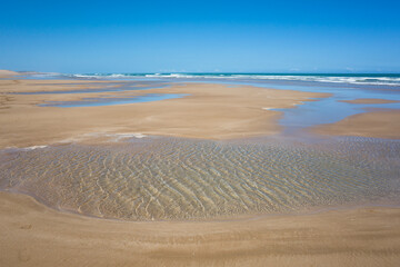 Low tide on the sandy beach of Essaouira, Morocco. A shallow water pool in the foreground with wind ripples on its surface, and reflections of the blue sky on wet sand