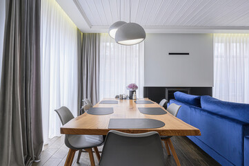 Modern dining room with a wooden table, gray chairs, and soft lighting. A blue velvet sofa adds a pop of color