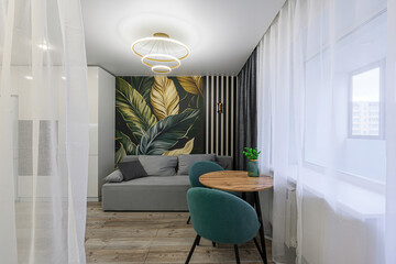 stylish room featuring a sofa, table with two chairs, and striking leaf-themed wallpaper. space is bathed in light from the window, softened by sheer curtains. modern light fixture hangs from ceiling