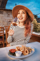 Young woman wearing a hat enjoying a glass of white wine and a plate of fried calamari with french fries in an outdoor restaurant