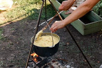 Outdoor cooking scene with a cauldron on a tripod over an open fire, one hand holding the pot and...