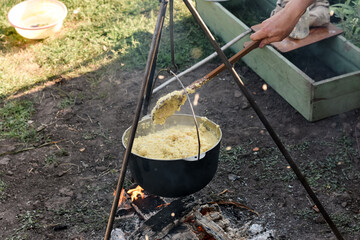 Cooking porridge in a cauldron over an open fire on a tripod, woman’s hand stirring with a wooden...
