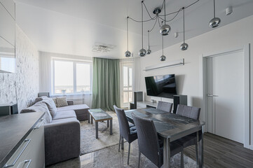 bright, modern living space featuring a gray sofa, dining set, and a large TV. Chrome light fixtures add a stylish touch