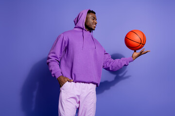 Stylish young man in casual purple hoodie spinning an orange basketball against a matching violet background