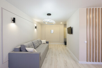 bright, modern living room features a gray sofa, light wood flooring, and minimalist décor. Wall sconces, a modern chandelier, and a TV provide lighting and entertainment