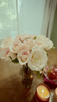 Close-up of a bouquet of pale pink and white roses in a glass vase on a wooden table, soft daylight from the window creates a warm romantic atmosphere, perfect for weddings or decor themes