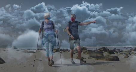 Walking couple using trekking poles and wearing face coverings, carrying backpack along tidal shore