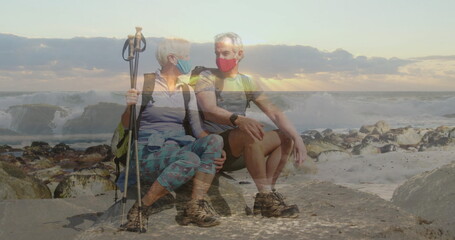 Sitting senior couple wearing hiking gear and masks on rocky shoreline, holding backpacks and poles