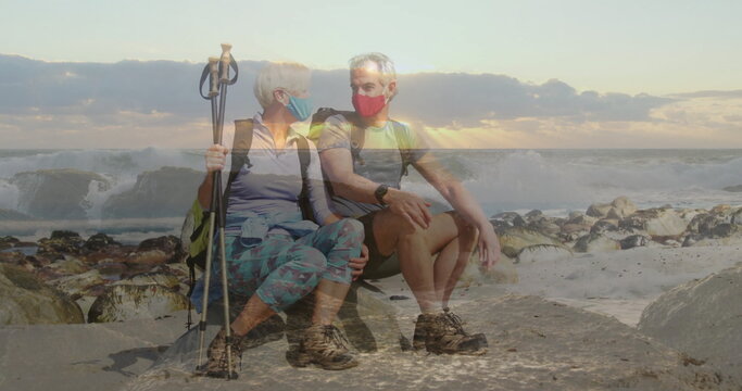 Sitting senior couple wearing hiking gear and masks on rocky shoreline, holding backpacks and poles - Powered by Adobe