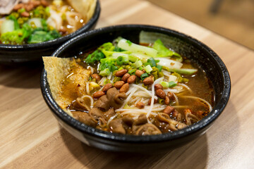 Bowl of spicy rice noodles with pork intestines, peanuts, green vegetables and chili broth served in black bowl on wooden dining table