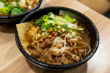 Close up of spicy rice noodles topped with pork intestines, peanuts, fresh greens and chili broth in black bowl, traditional Chinese noodle dish