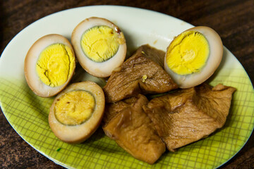 Assorted braised side dishes including soy marinated boiled eggs and tofu pieces, richly colored from soy sauce braising liquid, served on a patterned plate