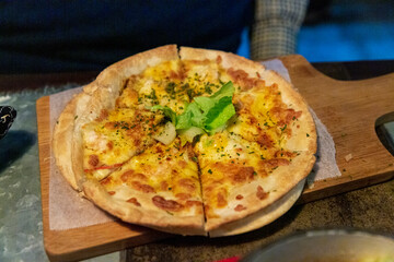Freshly baked cheese pizza served on wooden board, topped with melted mozzarella, herbs, and garnish, viewed in cozy dining setting