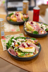 Fresh salad bowl with prosciutto, soft bun, feta cheese, beets, and leafy greens served with floral napkin and canned drinks on wood table