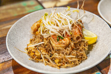Plate of Pad Thai noodles with prawns, tofu, bean sprouts, and lemon wedge served in ceramic bowl on wooden table in a cozy restaurant setting