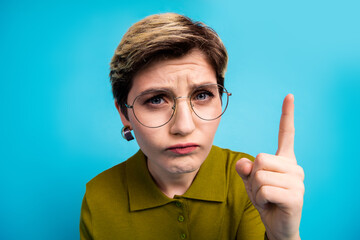 Confident young woman wearing khaki shirt pointing up against blue background while expressing an...