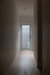 Narrow indoor hallway with wooden flooring leading to frosted glass door, softly lit with minimalist white walls and modern clean design