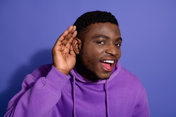 Young male expressing joy and holding hand to ear against purple background, wearing casual hoodie...