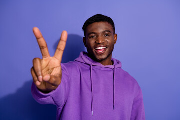 Young man wearing a purple hoodie making a v sign gesture while smiling with a purple background