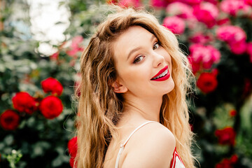 Beautiful blonde woman with a bright smile outdoors surrounded by vibrant red roses on a sunny day