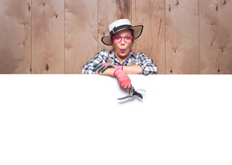 Surprised young gardener women behind white board with pruning shears on wooden background in...