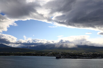 A panoramic view of Akureyri, the largest town in northern Iceland, captured from the opposite side of the fjord. The scene shows the coastal city with surrounding mountains, dramatic clouds