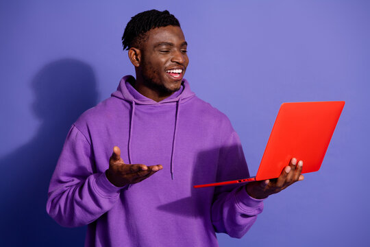 Young man in a purple hoodie holding a red laptop with a cheerful expression against a violet background - Powered by Adobe
