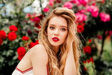 Attractive young blonde woman posing outdoors surrounded by vibrant red flowers under sunlight in...