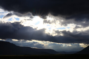A powerful sky scene with heavy dark clouds pierced by sharp rays of bright sunlight. The dramatic contrast between shadow and light creates a striking and atmospheric mood.